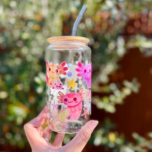 May include: A clear glass can-shaped drinking glass with a bamboo lid and a curved glass straw. The glass is decorated with colorful axolotl cartoon illustrations. The axolotls are pink, orange, and purple, with star and bubble accents.