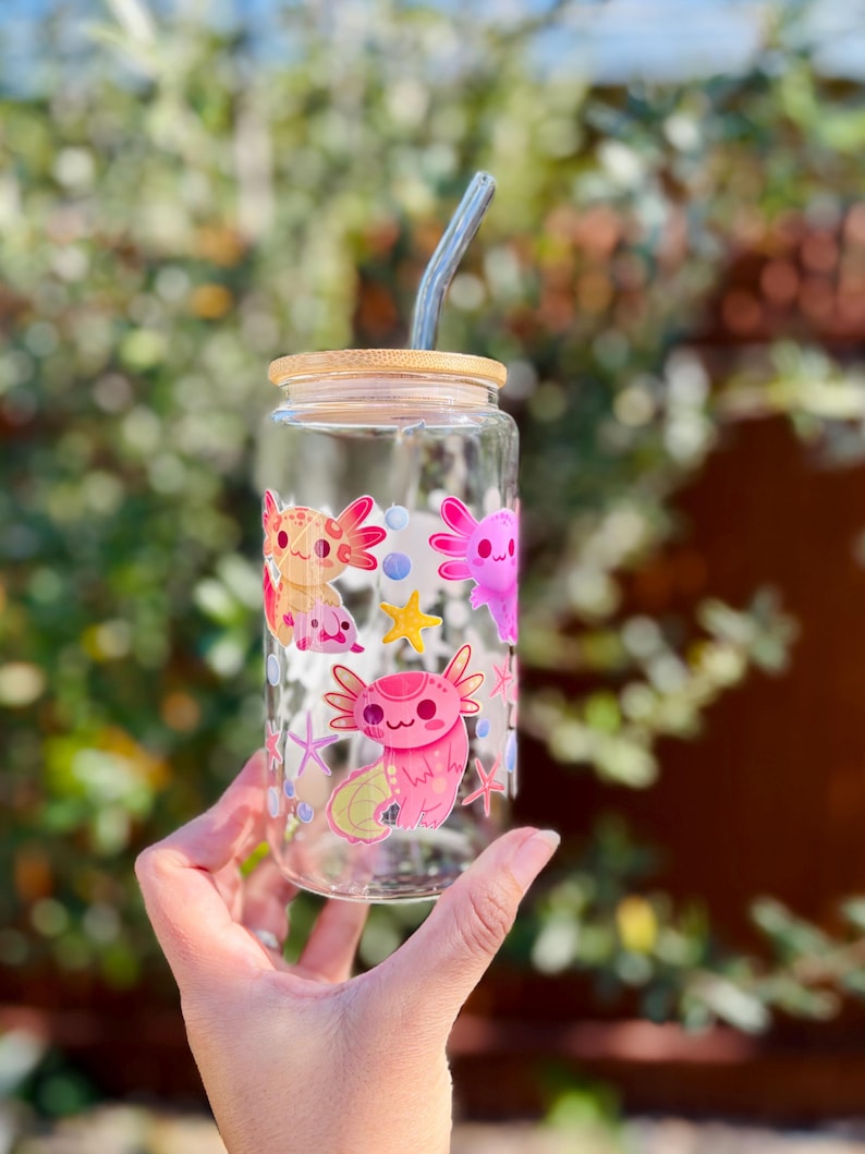 May include: A clear glass can-shaped drinking glass with a bamboo lid and a curved glass straw. The glass is decorated with colorful axolotl cartoon illustrations. The axolotls are pink, orange, and purple, with star and bubble accents.