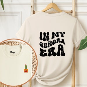 May include: A cream colored t-shirt with short sleeves and a black graphic print that says "In My Se&ntilde;ora Era" on the back. The shirt has a small cactus graphic on the front.