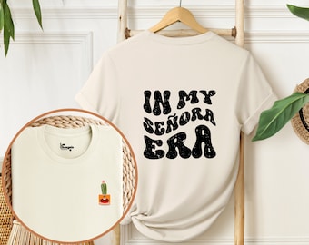 May include: A cream colored t-shirt with short sleeves and a black graphic print that says "In My Señora Era" on the back. The shirt has a small cactus graphic on the front.