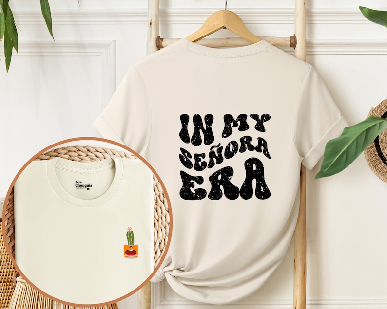 May include: A cream colored t-shirt with short sleeves and a black graphic print that says "In My Se&ntilde;ora Era" on the back. The shirt has a small cactus graphic on the front.
