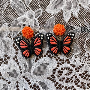 K&ouml;nnte beinhalten: Ein Paar schwarze Schmetterlingsohrringe mit orangefarbenen und wei&szlig;en Akzenten. Die Schmetterlinge haben einen Blumenanh&auml;nger oben.
