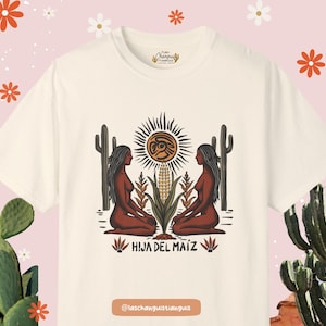 Op de afbeelding: Een wit T-shirt met een grafisch ontwerp met twee vrouwen die rug aan rug zitten voor een zon en ma&iuml;sstengels. De tekst "HIJA DEL MAIZ" is onder de afbeelding gedrukt. De tekst "Las Changuitas Tianguis" is in een kleiner lettertype bovenaan het shirt gedrukt. De tekst "@laschanguitastianguis" is onderaan het shirt gedrukt.
