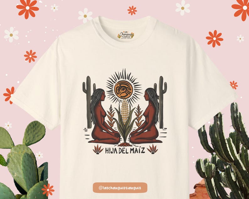 Op de afbeelding: Een wit T-shirt met een grafisch ontwerp met twee vrouwen die rug aan rug zitten voor een zon en ma&iuml;sstengels. De tekst "HIJA DEL MAIZ" is onder de afbeelding gedrukt. De tekst "Las Changuitas Tianguis" is in een kleiner lettertype bovenaan het shirt gedrukt. De tekst "@laschanguitastianguis" is onderaan het shirt gedrukt.