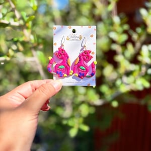 May include: A pair of pink cat-shaped earrings with yellow, blue, and green accents. The earrings are on a white card with the text "Las Changuis Tiancuis Handmade Jewelry". The earrings are held in front of a blurred green and brown background.