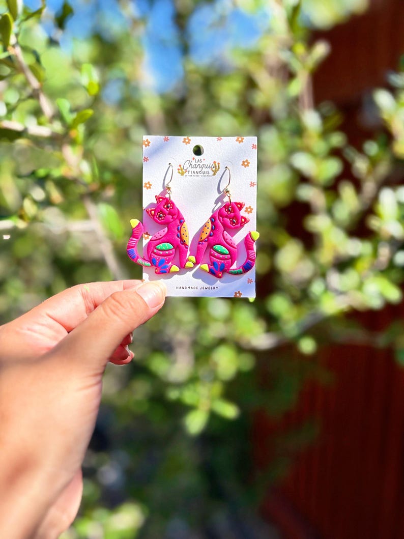 May include: A pair of pink cat-shaped earrings with yellow, blue, and green accents. The earrings are on a white card with the text "Las Changuis Tiancuis Handmade Jewelry". The earrings are held in front of a blurred green and brown background.