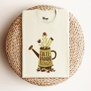 Puede incluir: Una camiseta blanca con una ilustración de una regadera verde y el texto "Culti-vando bilingües".
