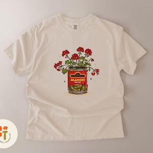 以下が含まれることがあります： クリーム色のTシャツ。ハラペーニョの缶から赤い花が咲いているグラフィック。缶には緑色のハラペーニョが詰まっています。「Jalapenos」の文字が太字でプリントされています。ユニークなデザインです。