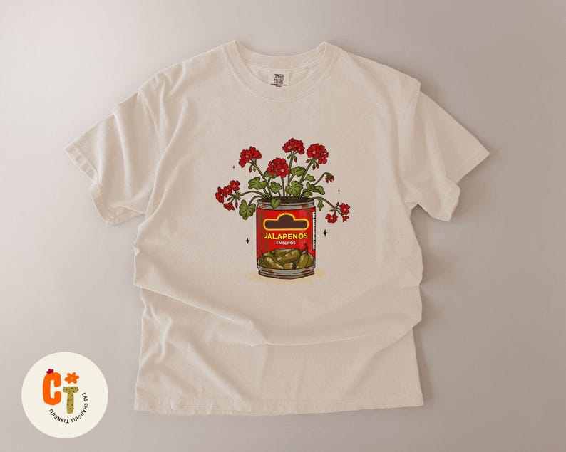 以下が含まれることがあります： クリーム色のTシャツ。ハラペーニョの缶から赤い花が咲いているグラフィック。缶には緑色のハラペーニョが詰まっています。「Jalapenos」の文字が太字でプリントされています。ユニークなデザインです。