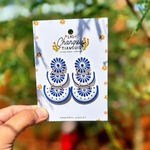May include: White and blue tiered earrings with a floral design are displayed on a white card. The card has the text "LAS Changuis TIANGUIS LATINA OWNED * HANDMADE" and "HANDMADE JEWELRY". The earrings are held by a hand.