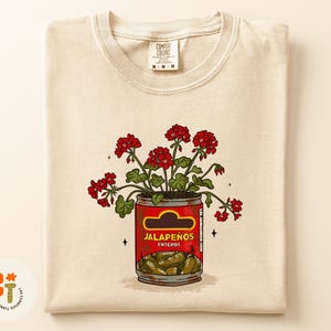 以下が含まれることがあります： クリーム色のTシャツ。赤い花が「Jalape&ntilde;os Enteros」の缶に入ったグラフィック。缶は赤色で、黄色のラベルと緑色のハラペーニョが入っています。シャツにはComfort Colorsのタグが付いています。