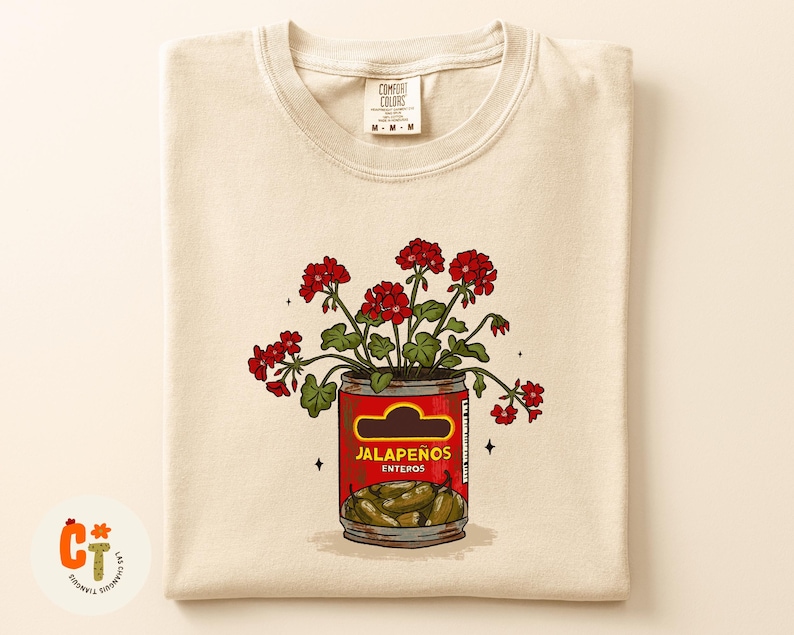 以下が含まれることがあります： クリーム色のTシャツ。赤い花が「Jalape&ntilde;os Enteros」の缶に入ったグラフィック。缶は赤色で、黄色のラベルと緑色のハラペーニョが入っています。シャツにはComfort Colorsのタグが付いています。