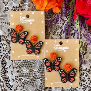 K&ouml;nnte beinhalten: Zwei Paar handgefertigte Schmetterlings-Ohrringe mit orangefarbenen Blumenakzenten. Die Ohrringe befinden sich auf braunen Karten mit dem Text "Las Changuis Tianguis" und "Handgefertigte Schmuckst&uuml;cke".