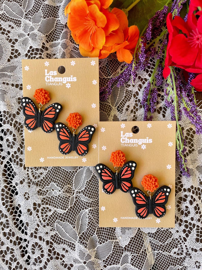 K&ouml;nnte beinhalten: Zwei Paar handgefertigte Schmetterlings-Ohrringe mit orangefarbenen Blumenakzenten. Die Ohrringe befinden sich auf braunen Karten mit dem Text "Las Changuis Tianguis" und "Handgefertigte Schmuckst&uuml;cke".
