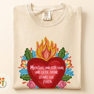 Peut inclure: T-shirt crème avec un motif coloré. Le design présente un cœur rouge avec des flammes, entouré de fleurs et de feuilles. Le texte espagnol dit "Mientras uno está vivo, uno debe amar lo más que pueda".