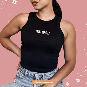 K&ouml;nnte beinhalten: Schwarzes geripptes Tanktop mit dem Text "ya wey" in gotischer Schrift. Das Oberteil ist &auml;rmellos und hat einen runden Ausschnitt. Das Model tr&auml;gt eine blaue Jeans. Der Hintergrund ist rosa mit wei&szlig;en Blumengrafiken.