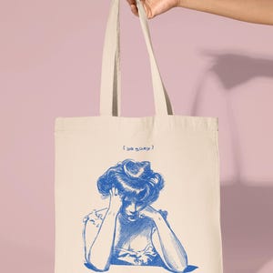 May include: A white canvas tote bag with blue text that reads "[ya quey]" and a blue illustration of a woman with her head in her hands.