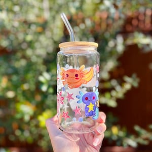 May include: A clear glass tumbler with a bamboo lid and a glass straw. The tumbler is decorated with colorful cartoon axolotls, starfish, and bubbles. The axolotls are orange and blue. The tumbler is held by a person.