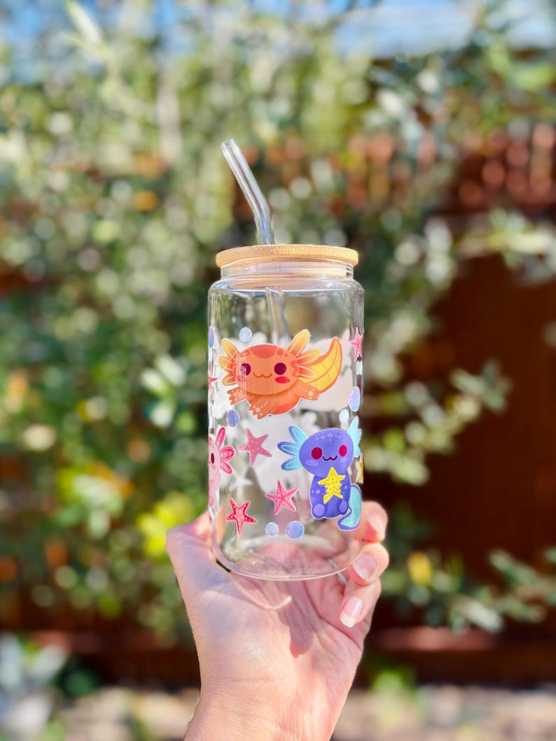 May include: A clear glass tumbler with a bamboo lid and a glass straw. The tumbler is decorated with colorful cartoon axolotls, starfish, and bubbles. The axolotls are orange and blue. The tumbler is held by a person.