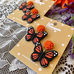 K&ouml;nnte beinhalten: Ein Paar handgefertigte Ohrringe mit schwarzen und orangefarbenen Schmetterlingsanh&auml;ngern und kleinen orangefarbenen Blumenakzenten. Die Ohrringe werden auf einer braunen Karte mit dem Text "Las Changuis Tianguis" und "Handmade Jewelry" pr&auml;sentiert.