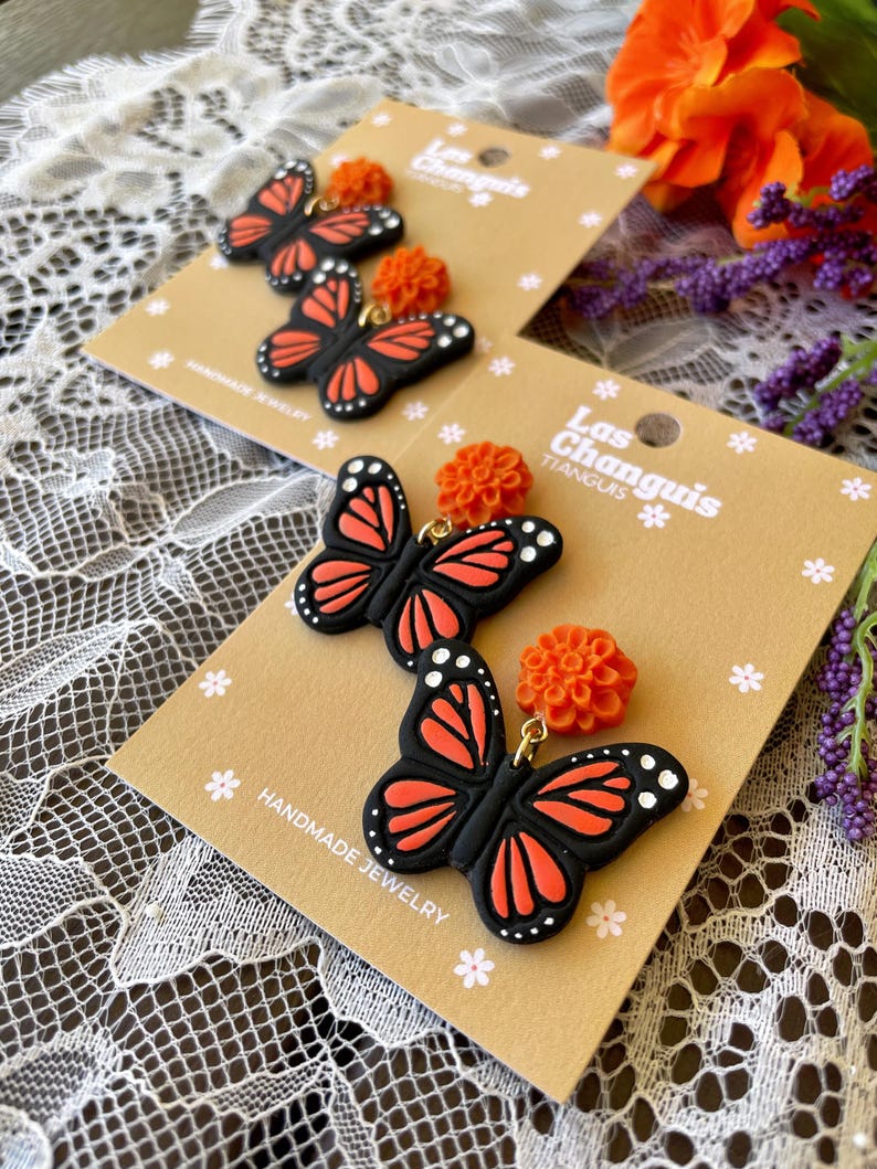 K&ouml;nnte beinhalten: Ein Paar handgefertigte Ohrringe mit schwarzen und orangefarbenen Schmetterlingsanh&auml;ngern und kleinen orangefarbenen Blumenakzenten. Die Ohrringe werden auf einer braunen Karte mit dem Text "Las Changuis Tianguis" und "Handmade Jewelry" pr&auml;sentiert.