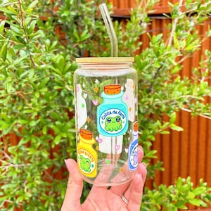 May include: A clear glass bottle with a bamboo lid and a straw. The bottle has colorful stickers with frogs and the text "Collita de Rana".