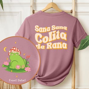 Może przedstawiać: Koszulka w kolorze lila z napisem "Sana Sana Colita de Rana" w retro stylu. Z przodu koszulki znajduje się kresk&oacute;wkowa żaba w kapeluszu z grzyba, siedząca na lilii wodnej.