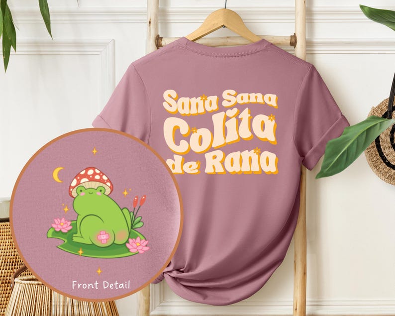 Może przedstawiać: Koszulka w kolorze lila z napisem "Sana Sana Colita de Rana" w retro stylu. Z przodu koszulki znajduje się kresk&oacute;wkowa żaba w kapeluszu z grzyba, siedząca na lilii wodnej.