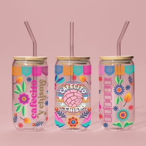 May include: Three clear glass tumblers with colorful floral and geometric designs. Each tumbler has a bamboo lid and a reusable straw. The tumblers feature the text "Cafecito y chisme" and "Cafecito y chisme".