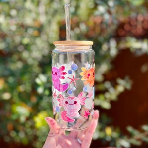 May include: A clear glass can-shaped drinking glass with a bamboo lid and a clear straw. The glass is decorated with pink and orange axolotl illustrations, starfish, and bubbles. The glass is held in a hand against a blurred green background.