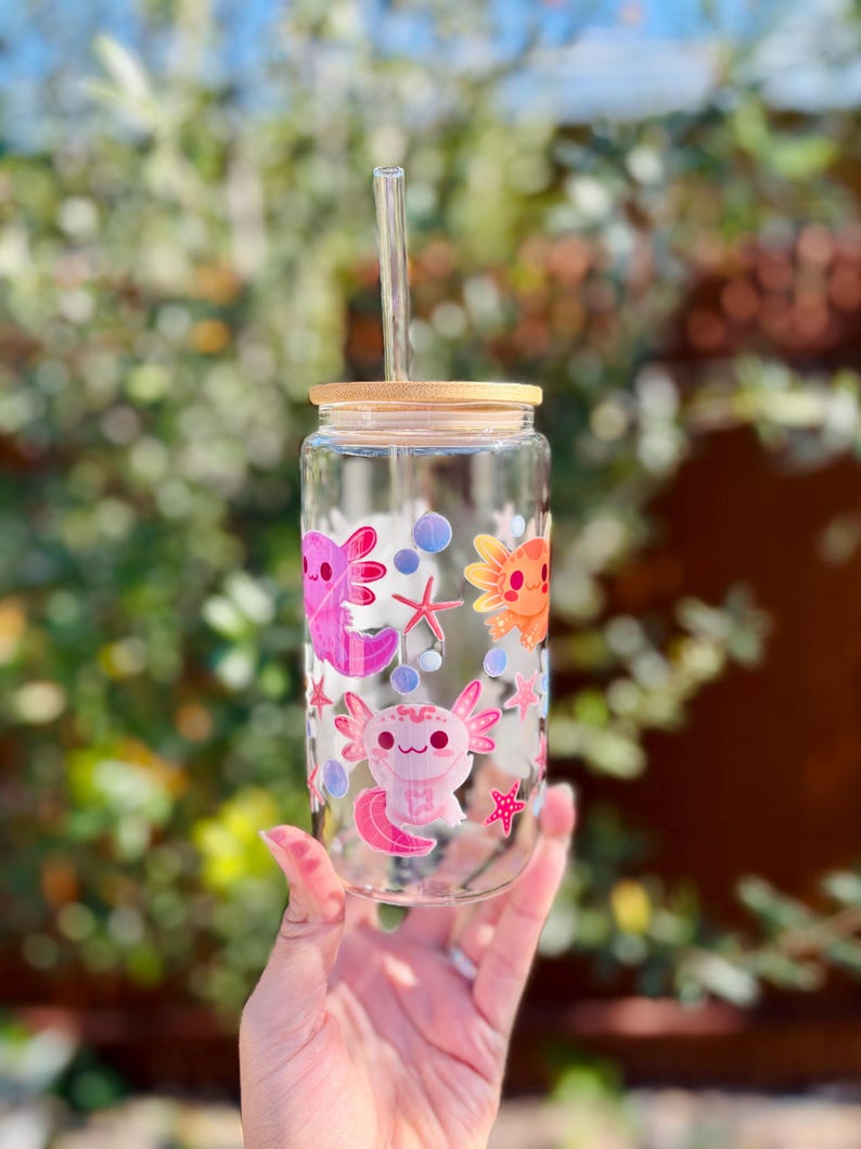 May include: A clear glass can-shaped drinking glass with a bamboo lid and a clear straw. The glass is decorated with pink and orange axolotl illustrations, starfish, and bubbles. The glass is held in a hand against a blurred green background.