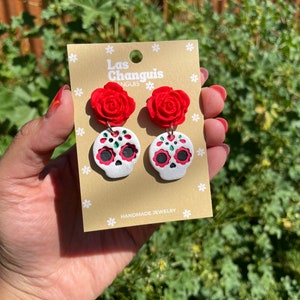 May include: A pair of handmade sugar skull earrings with red roses. The earrings are on a card that says "Las Changuis" and "Handmade Jewelry".