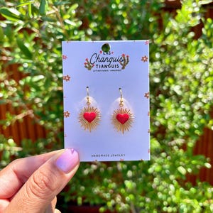 May include: Gold heart earrings with red enamel and a sunburst design. The earrings are on a white card with the text "Las Changuis Tianguis Latina Owned Handmade Jewelry".