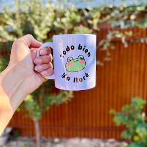 May include: White ceramic mug featuring a cartoon frog with the phrase "Todo bien ya llor&eacute;" in black script. The frog is green with pink cheeks. The mug is held against a blurred outdoor background.