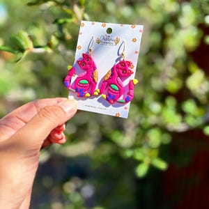 May include: A pair of handmade pink cat earrings with gold-colored hooks. The earrings feature colorful details in yellow, orange, blue, and green. The earrings are displayed on a white card with the text "LAS CHARQUIS TIANGUIS HANDMADE JEWELRY".