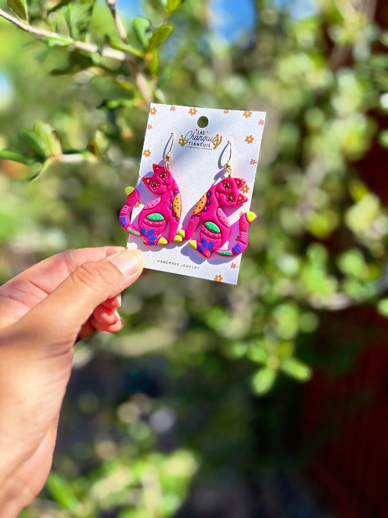 May include: A pair of handmade pink cat earrings with gold-colored hooks. The earrings feature colorful details in yellow, orange, blue, and green. The earrings are displayed on a white card with the text "LAS CHARQUIS TIANGUIS HANDMADE JEWELRY".