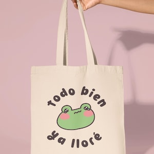 May include: A beige canvas tote bag with a green frog cartoon illustration and the text "todo bien ya llor&eacute;".