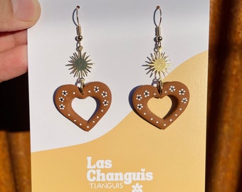 May include: A pair of brown heart-shaped earrings with white flower accents and gold sun charms. The earrings are on a white card with the text "Las Changuis Tianguis laschanguistianguis.shop".