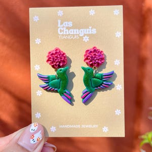 May include: A pair of handmade green hummingbird earrings with pink flower accents. The earrings are on a card that says "Las Changuis Tianguis Handmade Jewelry".