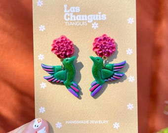 Könnte beinhalten: Ein Paar handgefertigte grüne Kolibri-Ohrringe mit rosa Blumenakzenten. Die Ohrringe befinden sich auf einer Karte mit der Aufschrift "Las Changuis Tianguis Handmade Jewelry".