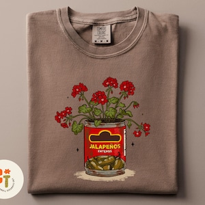 以下が含まれることがあります： 赤唐辛子の缶と赤い花のグラフィックが描かれた茶色のTシャツ。「Jalape&ntilde;os Enteros」の文字とハラペーニョの写真が描かれた赤い缶。TシャツにはComfort Colorsのタグが付いています。