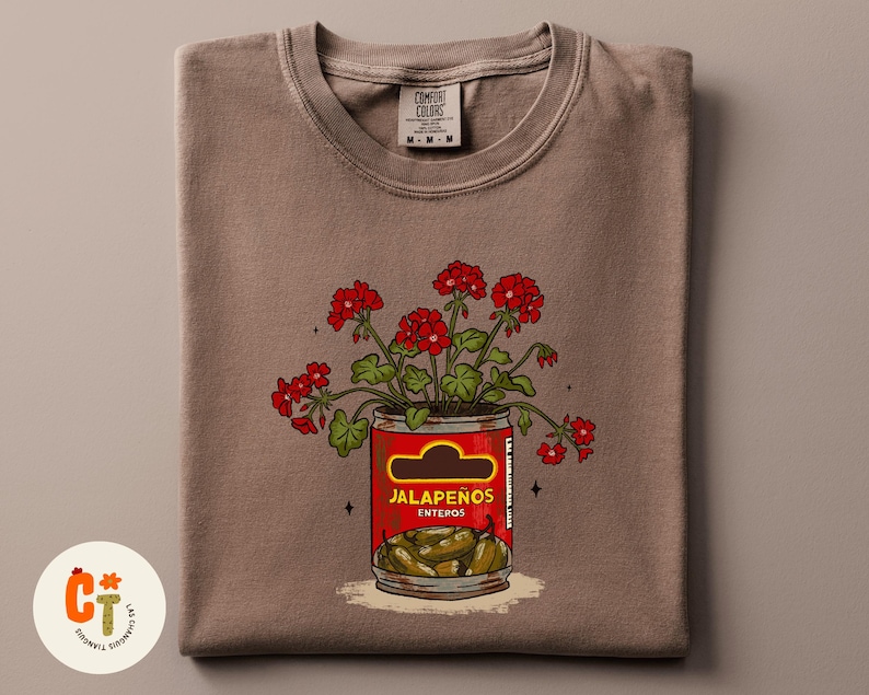 以下が含まれることがあります： 赤唐辛子の缶と赤い花のグラフィックが描かれた茶色のTシャツ。「Jalape&ntilde;os Enteros」の文字とハラペーニョの写真が描かれた赤い缶。TシャツにはComfort Colorsのタグが付いています。