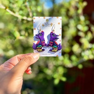 May include: A pair of handmade purple bird earrings with gold hooks. The earrings feature colorful details in red, green, and yellow. The earrings are displayed on a white card with the text "Las Changuis Tianguis Handmade Jewelry".