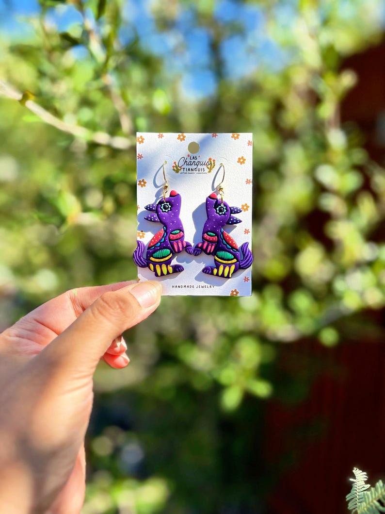 May include: A pair of handmade purple bird earrings with gold hooks. The earrings feature colorful details in red, green, and yellow. The earrings are displayed on a white card with the text "Las Changuis Tianguis Handmade Jewelry".