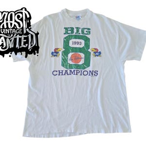 以下が含まれることがあります： 緑と青のグラフィックが特徴の白いヴィンテージTシャツ。「BIG 8 1993 CHAMPIONS」の文字とバスケットボール、ジェイホークのマスコットが描かれています。シャツには、白黒のグラフィティスタイルで「MOST VINTAGE WANTED」の文字も入っています。