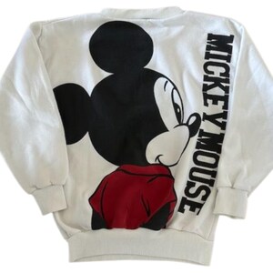 May include: White sweatshirt featuring a large Mickey Mouse graphic on the back. The design includes a black Mickey Mouse head, a red shirt, and the words "Mickey Mouse" in black block letters. The sweatshirt has long sleeves.