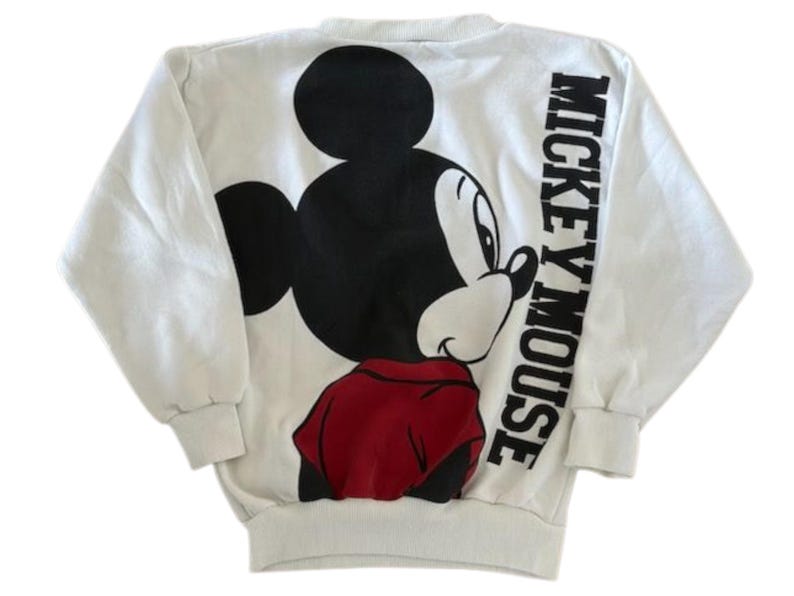 May include: White sweatshirt featuring a large Mickey Mouse graphic on the back. The design includes a black Mickey Mouse head, a red shirt, and the words "Mickey Mouse" in black block letters. The sweatshirt has long sleeves.
