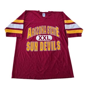 May include: A maroon V-neck jersey with short sleeves featuring yellow and white stripes. The front displays the text ARIZONA STATE in yellow, a white oval with XXL, and the text SUN DEVILS in yellow. The shirt has a white tag at the neckline.