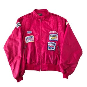 May include: A bright pink racing jacket featuring a stand-up collar, long sleeves, and a front zip fastening. The jacket is decorated with various embroidered patches, including NHRA, Winston Drag Racing, and U.S. Nationals logos with text.