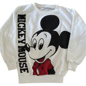May include: White sweatshirt featuring a large graphic of Mickey Mouse in black, red, and white. The words "MICKEY MOUSE" are printed vertically on the left sleeve. The sweatshirt has a crew neck and long sleeves.
